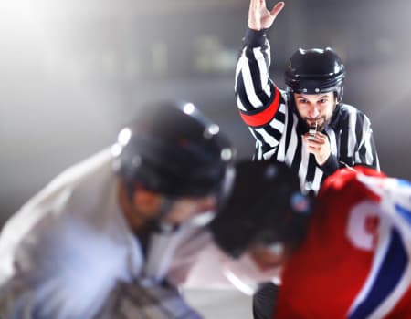 Ice Hockey Referee with hand up at face off of a power play.  Two players lean towards the puck drop